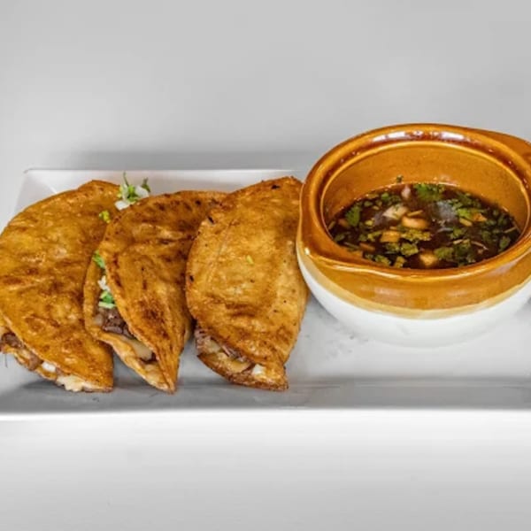 Three golden-brown La Calera birria tacos with a side of rich consommé on a white rectangular plate.